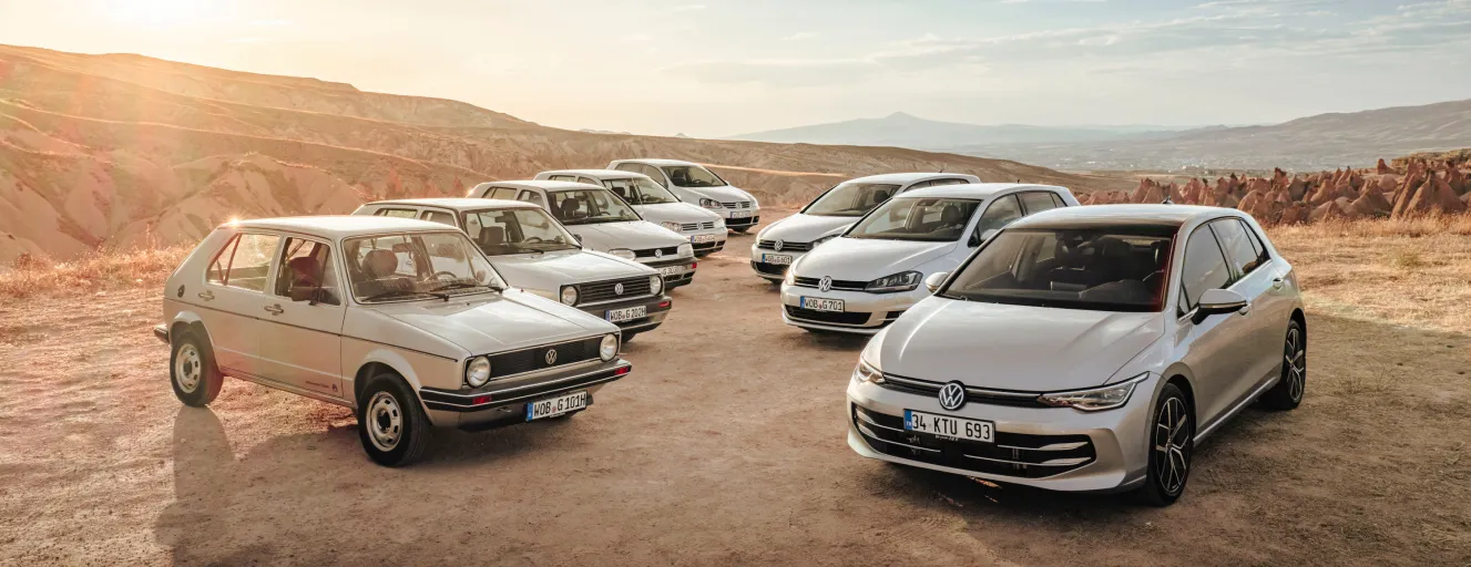 A group of white Volkswagen cars, featuring vintage models and the modern Tayron, parked together on a desert-style landscape.